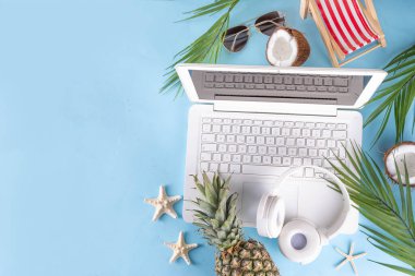 İş ve tatil. Yaratıcı yaz ofis dairesi beyaz dizüstü bilgisayarı, tropikal yaz aksesuarları, palmiye yaprakları, güneş gözlükleri, parmak arası terlikler, ananas ve açık mavi arka planda hindistan cevizi ile yatıyordu.