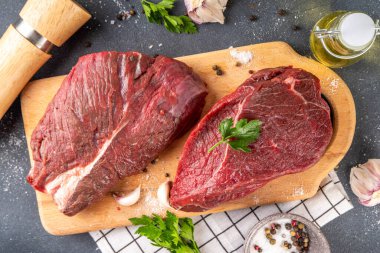 Çiğ biftekler ızgara yapmaya hazır. Kesme tahtasında iki biftek, otlar, baharatlar, zeytinyağı, yemek masasının üstünde fotokopi alanı.