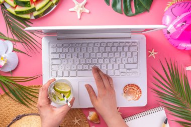 Yazlık ofis, çalışma geçmişi, blogcu iş yeri düzlüğü. Not defteri, hasır şapka, tropikal dekor ve yapraklarla dolu beyaz dizüstü bilgisayar klavyesi, açık pembe arka planda soğuk kokteyl bardağı.