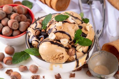 Fındıklı çikolatalı dondurma. Fındıklı dondurma kepçeli büyük kase porsiyonu, çikolata sosu ve mutfak masasının arkasında bir sürü fındık. 