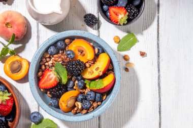 Meyve ve böğürtlenli granola kahvaltısı, yaz tatili lezzetli kahvaltı kasesi bol meyve ve böğürtlenli, süt testisi beyaz ahşap masada.