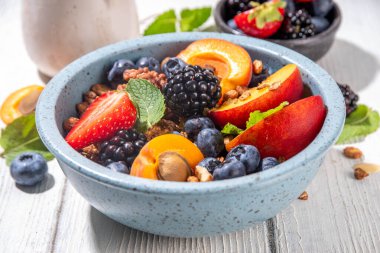 Meyve ve böğürtlenli granola kahvaltısı, yaz tatili lezzetli kahvaltı kasesi bol meyve ve böğürtlenli, süt testisi beyaz ahşap masada.