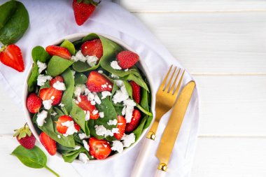 Ispanak ve çilekli yaz salatası süzme peynir ve balzamik sos taze çiğ sebze kasesi sağlıklı diyet yemeği beyaz mutfak masasında çatal bıçak takımı.