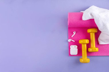 Spor ve spor geçmişi. Kadın spor aksesuarları. Halterli yoga minderi, su şişesi, havlu, ip atlama, kulaklık açık mor arka planda, boşluk kopyala