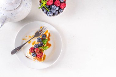 Geleneksel New York peynirli kek dilimleri tabakta, taze böğürtlenler beyaz mutfak masasında