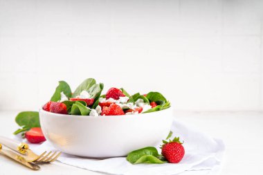 Ispanak ve çilekli yaz salatası süzme peynir ve balzamik sos taze çiğ sebze kasesi sağlıklı diyet yemeği beyaz mutfak masasında çatal bıçak takımı.