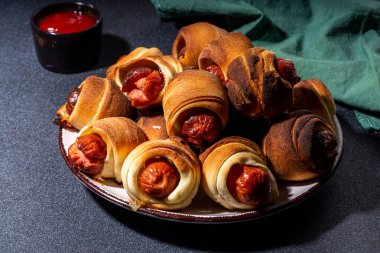 Traditional Christmas dish recipe Pigs in blankets, sausages wrapped in bacon, oven baked puff pastry hot dogs rolls with xmas tree branches, decoration and gifts on black table 