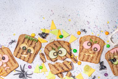 Funny halloween monster sandwiches. Set of various decorated monster sandwich and toasts, with cucumber, cheese and meat sausages, white table background with Halloween decorations