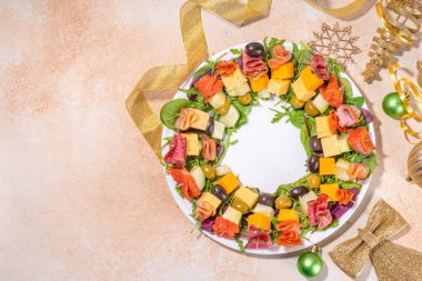 Noel partisi yemeği, modaya uygun mezeler. Sosis, et, peynir, zeytin, bej masa arkaplanındaki yeşillikler ve altın rengi Noel dekorasyonundan yapılmış çelenk.
