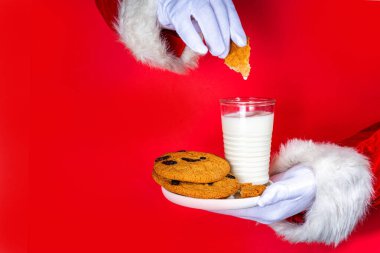 Noel Baba 'nın eli, geleneksel Noel kurabiyesi atıştırmalığıyla süt bardağıyla. Noel Baba Batıran Kurabiye, parlak kırmızı yırtık kağıt delikli arka planda. Mutlu Noeller ve mutlu yıllar tebrik kartı.