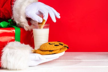Noel Baba 'nın eli, geleneksel Noel kurabiyesi atıştırmalığıyla süt bardağıyla. Santa Dunking Cookie, parlak kırmızı ve beyaz ahşap arka planda. Mutlu Noeller ve mutlu yıllar tebrik kartı.