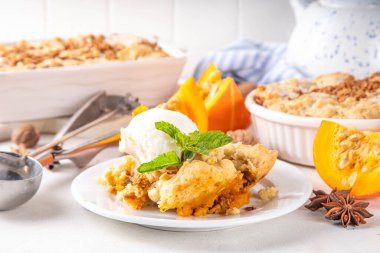 Homemade autumn pumpkin crisp cobbler, pumpkin crumble pie with crunchy dough topping in baking dish and plate, with ice cream scoop on white kitchen table clipart