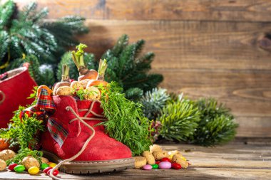 Sinterklaas, Saint Nicholas day background. Little kid red shoe with carrots and sweets, trooigoed, pepernoten, candy. Dutch Christmas holiday on cozy wooden background. 