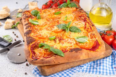 Domates ve fesleğenli ev yapımı margarita pizza, malzemeler ve arka planda beyaz mutfak masasında zeytinyağı.