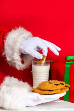 Noel Baba 'nın eli, geleneksel Noel kurabiyesi atıştırmalığıyla süt bardağıyla. Santa Dunking Cookie, parlak kırmızı ve beyaz ahşap arka planda. Mutlu Noeller ve mutlu yıllar tebrik kartı.