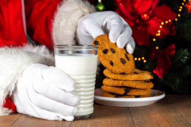 Noel Baba kurabiye ve süt yiyor. Noel Baba elleri Noel ağacına karşı ahşap masada kurabiye ve süt bardağı topluyor., 