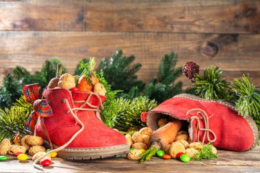Sinterklaas, Saint Nicholas day background. Little kid red shoe with carrots and sweets, trooigoed, pepernoten, candy. Dutch Christmas holiday on cozy wooden background. 