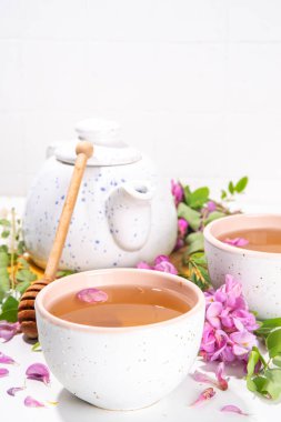 Organik bitkisel sağlık içeceği. Akasya çiçeği yeşili çay, beyaz fincanlarda, çaydanlıkla, beyaz bir masanın üstünde, kopyalanacak yer ve çiçek açan akasya çiçekleri.