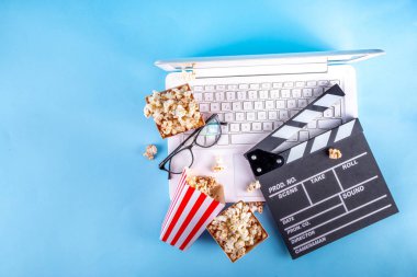 Sinema, film ve film izleme gecesi konsepti. Alkış tahtası, çizgili patlamış mısır kovası kutuları, açık mavi arka planda gözlüklü ve beyaz dizüstü bilgisayarlı, boşluğu kopyala