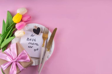 Anneler günü sofrası kurulumu. Pembe Anneler Günü 'nde kahvaltı, öğle yemeği, yemek menüsü, davetiye maketi. Altın çatal bıçak, tabak, lale çiçeği ve hediye kutusuyla güzel bir masa.