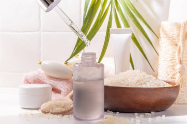 Pirinç suyu. Doğal organik pirinç kozmetiği. Mayalanmış güzellik, sağlık ve kaplıca. Şeffaf beyaz serum, beyaz banyo arka planında pirinç taneleri ve banyo malzemeleriyle şişe düşürür.