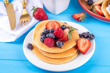 Yaz meyveli geleneksel Amerikan krepleri yığını. Sabah güneşinde bal, çilek ve yabanmersinli kahvaltı krepleri, mavi ahşap arka plan fotokopi alanı.