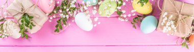 Pink Easter greeting card background with spring flowers, green leaf branches and colorful pastel Easter eggs, top view copy space