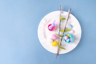 Paskalya Bayramı menü seti, pastel mavi arka planda çatal bıçak takımı ve tabak kareli peçete, şenlikli kurdele, şirin tavşanlı Paskalya bayramı dekoru, paskalya çikolatası yumurtaları.