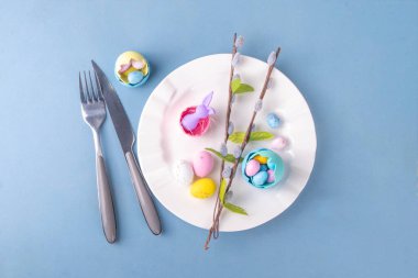Paskalya Bayramı menü seti, pastel mavi arka planda çatal bıçak takımı ve tabak kareli peçete, şenlikli kurdele, şirin tavşanlı Paskalya bayramı dekoru, paskalya çikolatası yumurtaları.