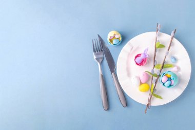 Paskalya Bayramı menü seti, pastel mavi arka planda çatal bıçak takımı ve tabak kareli peçete, şenlikli kurdele, şirin tavşanlı Paskalya bayramı dekoru, paskalya çikolatası yumurtaları.