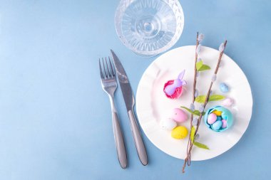 Paskalya Bayramı menü seti, pastel mavi arka planda çatal bıçak takımı ve tabak kareli peçete, şenlikli kurdele, şirin tavşanlı Paskalya bayramı dekoru, paskalya çikolatası yumurtaları.