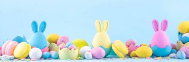 Tatlı ve parlak pastel renkli Paskalya yumurtaları ve tavşan arka planı. Pembe. Mavi, sarı renkli tavşanlar, yuvalarda ve yumurta kabuklarında çikolata yumurtaları, açık mavi arkaplanda şeker parçacıkları