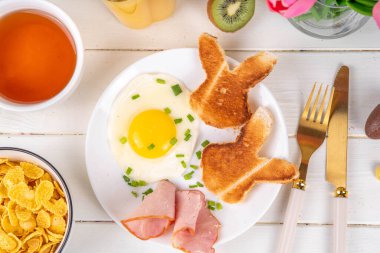 Paskalya kahvaltısı ya da kızarmış yumurta, pastırma dilimleri ve tavşancık suratı şeklinde kızarmış ekmek, bahar çiçekleri ve beyaz tahta masa üzerinde paskalya çikolatası yumurtaları.