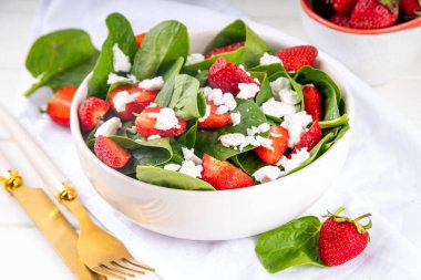 Ispanak ve çilekli yaz salatası süzme peynir ve balzamik sos taze çiğ sebze kasesi sağlıklı diyet yemeği beyaz mutfak masasında çatal bıçak takımı.
