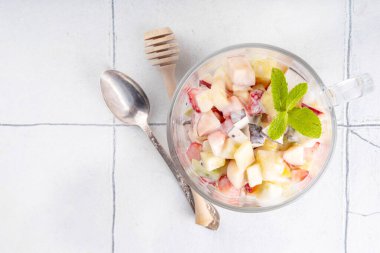 Elma, ananas, kivi, şeftali, çilek, muz, bal ve Yunan yoğurdu soslu kremalı yaz meyve salatası. Sağlıklı yaz diyeti yemek tarifi, beyaz mutfak masası fotokopi alanı üzerinde.