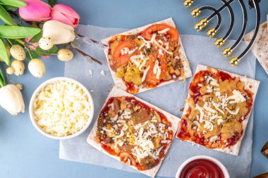 Açık mavi arkaplanda, içinde malzemeler, peynir ve domates sosu olan Matzah Pizza. Yahudi Pesach ve Hamursuz Bayramı için gıda fikirleri ve tarifi