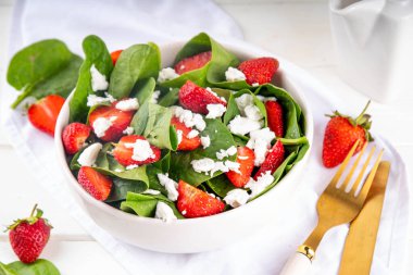 Ispanak ve çilekli yaz salatası süzme peynir ve balzamik sos taze çiğ sebze kasesi sağlıklı diyet yemeği beyaz mutfak masasında çatal bıçak takımı.