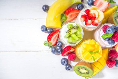 Cam kavanozlarda çeşitli meyve ve meyveli yoğurt seti. Çeşitli sağlıklı kahvaltılık yoğurt, yabanmersini, çilek, mango, kivi, ahududu taze meyve ve böğürtlen, beyaz ahşap arka plan