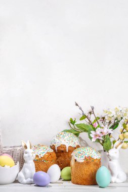 Şekerli Paskalya pastaları, renkli yumurta sepeti, seramik tavşanlar ve beyaz mutfak masasında bahar çiçekleri, fotokopi alanı. Mutlu Paskalya Tatilleri tebrik kartı arkaplan kopyalama alanı