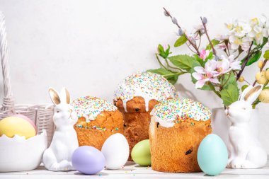 Şekerli Paskalya pastaları, renkli yumurta sepeti, seramik tavşanlar ve beyaz mutfak masasında bahar çiçekleri, fotokopi alanı. Mutlu Paskalya Tatilleri tebrik kartı arkaplan kopyalama alanı