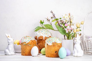 Şekerli Paskalya pastaları, renkli yumurta sepeti, seramik tavşanlar ve beyaz mutfak masasında bahar çiçekleri, fotokopi alanı. Mutlu Paskalya Tatilleri tebrik kartı arkaplan kopyalama alanı