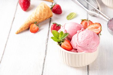 Yaz ferahlatıcı soğuk tatlı, lezzetli ve aromatik çilekli dondurma, seramik kasede ev yapımı meyve şerbeti, taze çilekler, beyaz ahşap arka planda dondurma külahları.