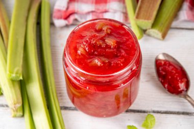 Pembe ve yeşil rengarenk tatlı reçel. Ev yapımı tatlı ravent konserveleri, beyaz ahşap masada küçük bir kavanozda, arka planda taze ravent ile.