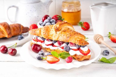 Krem şantili, taze meyve ve meyveli tatlı kahvaltı kruvasan sandviçi. Lezzetli bir yaz yemeği ya da içinde çilek, yabanmersini, kiraz, ahududu ve fındık bulunan taze kruvasanla atıştırmalık bir şeyler.