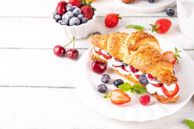 Krem şantili, taze meyve ve meyveli tatlı kahvaltı kruvasan sandviçi. Lezzetli bir yaz yemeği ya da içinde çilek, yabanmersini, kiraz, ahududu ve fındık bulunan taze kruvasanla atıştırmalık bir şeyler.