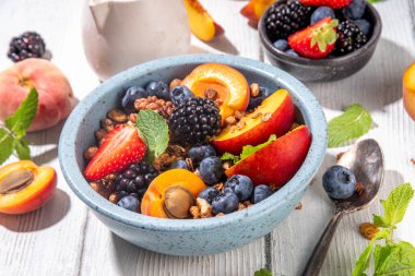 Meyve ve böğürtlenli granola kahvaltısı, yaz tatili lezzetli kahvaltı kasesi bol meyve ve böğürtlenli, süt testisi beyaz ahşap masada.