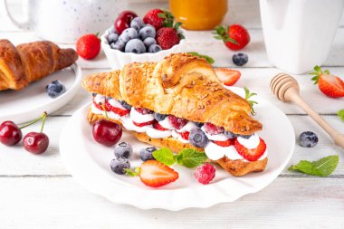 Krem şantili, taze meyve ve meyveli tatlı kahvaltı kruvasan sandviçi. Lezzetli bir yaz yemeği ya da içinde çilek, yabanmersini, kiraz, ahududu ve fındık bulunan taze kruvasanla atıştırmalık bir şeyler.