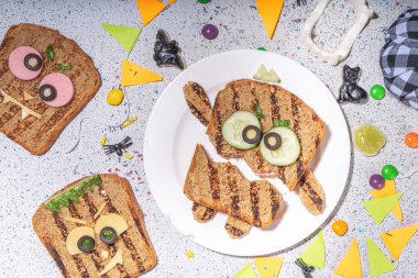 Funny halloween monster sandwiches. Set of various decorated monster sandwich and toasts, with cucumber, cheese and meat sausages, white table background with Halloween decorations