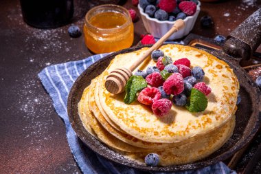 Homemade french tiny pancakes, battercakes with berry. Stack of freshly baked thin crepes on frying pan with fresh blueberry, raspberries and honey. Healthy morning breakfast on dark background