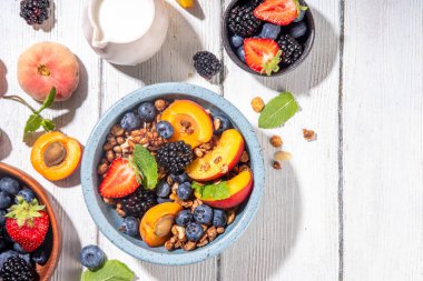 Meyve ve böğürtlenli granola kahvaltısı, yaz tatili lezzetli kahvaltı kasesi bol meyve ve böğürtlenli, süt testisi beyaz ahşap masada.